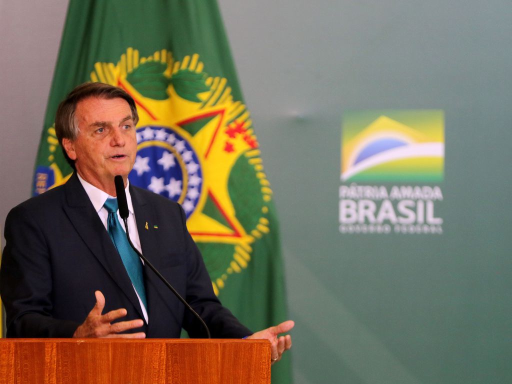 O Presidente da República Jair Bolsonaro durante evento no Palácio do Planalto; ele veste um terno preto e posa em frente a bandeira do Brasil