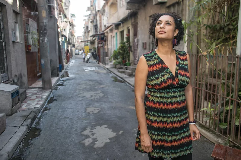 Marielle Franco era uma mulher negra com olhos e cabelos castanhos; na foto ela está usando um vestido colorido, atrás dela está uma rua de uma favela do Rio de Janeiro