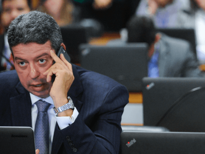 Arthur Lira é um homem branco com cabelos grisalhos; ele veste um terno preto e está com o telefone no ouvido