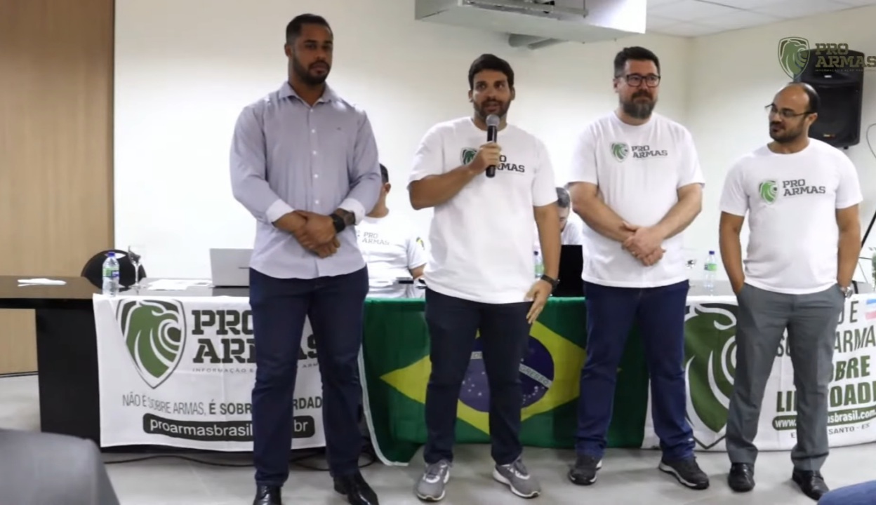 André Porciuncula é um homem branco com cabelos castanhos. Ele veste camiseta branca escrito "pró-armas" e segura um microfone na mão. Na imagem ele está discursando sobre o uso de recursos da Lei Rouanet para produção de conteúdos armamentistas