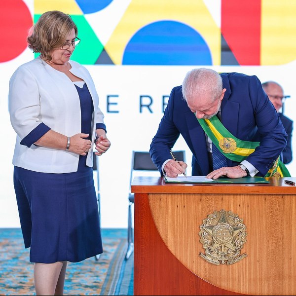 Cida Gonçalves durante posse como Ministra da Mulher. À esquerda, Cida é uma mulher branca com cabelos loiros e curtos. Ela veste um terno branco sobre um vestido azul marinho. Ao seu lado está o presidente Lula, assinando um documento apoiado em uma mesa com o brasão do Brasil. Lula veste terno azul e a faixa presidencial nas cores verde e amarelo. Lula é um homem branco com cabelos grisalhos.