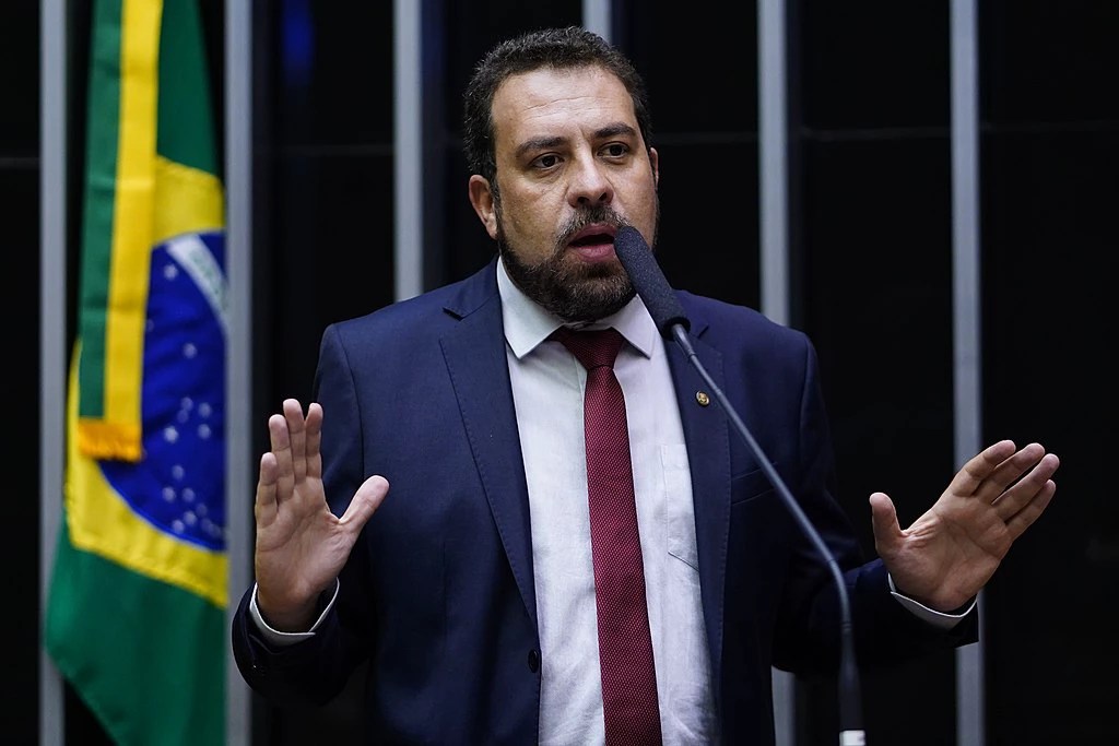 Deputado Guilherme Boulos é um homem branco com cabelos, olhos e barba castanhos. Ele veste terno azul marinho com gravata vermelha e camisa social branca. Boulos está falando ao microfone, com a bandeira do Brasil atrás