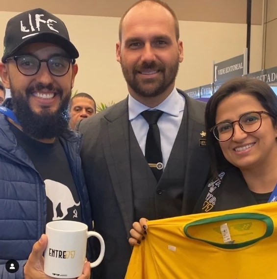 À esquerda Wilker Delkerson Amaral, um homem negro de barba, na imagem ele usa boné e óculos, e segura uma xícara, ao meio Eduardo Bolsonaro usando terno cinza e gravata preta, e à direita, Denise uma mulher branca com cabelos pretos, na imagem ela usa óculos e segura uma camiseta do Brasil.