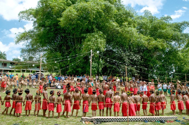 Indígenas durante festa tradicional do Turé. Na imagem é possível ver dezenas de indígenas, homens, mulheres e crianças, de mãos dadas em um círculo. Eles vestem roupas vermelhas e adereços tradicionais